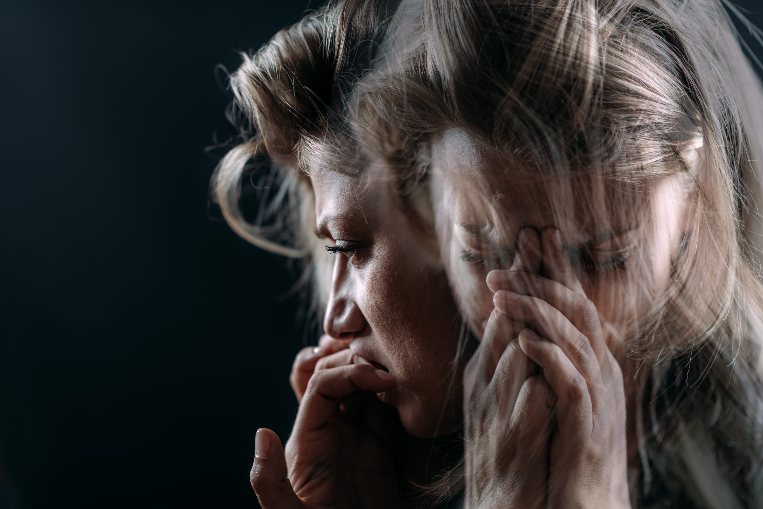 A blurred double exposure of a woman with light brown hair holding her head in distress reflects the urgency for trauma treatment. Her face shows signs of anxiety, and her hands cover parts of her face. The dark background emphasizes her emotional state, echoing the atmosphere of a trauma clinic.