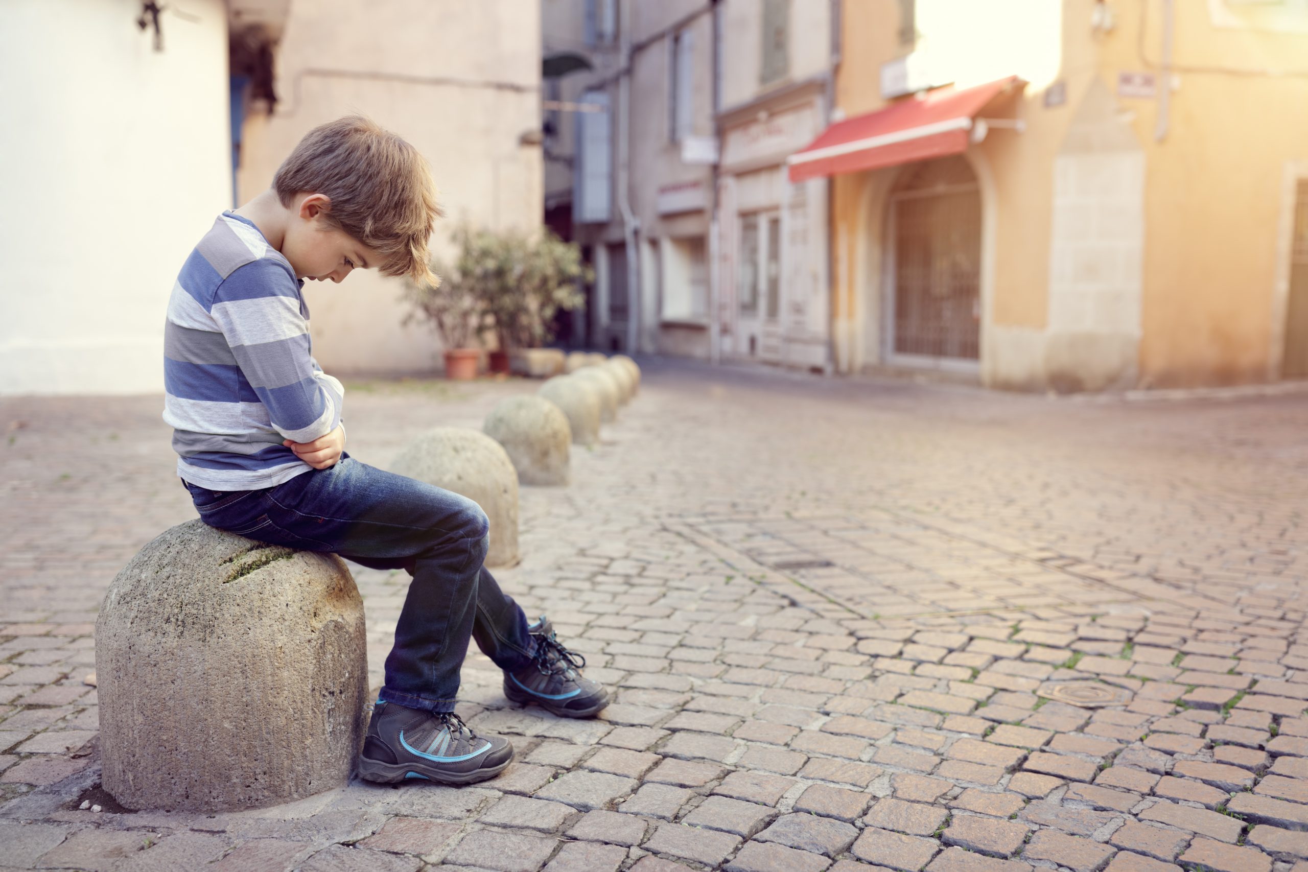 A young boy sits alone on a stone bollard in the middle of a quiet cobblestone street. He is wearing a striped shirt and jeans, and appears to be looking down, with his arms crossed. The buildings around him are old with pastel-colored facades, reminiscent of scenes near a trauma clinic or mental hospital.