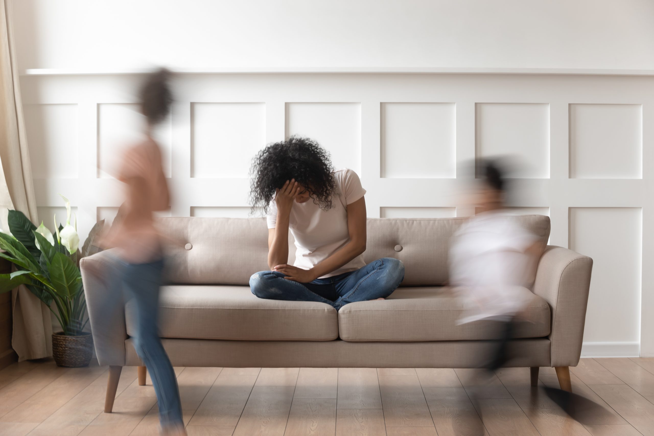 A person sits on a beige couch with their head in their hand, looking distressed. Two children are seen in motion, one on each side of the couch, creating a blur as they run by. The background shows a white paneled wall and a plant in the corner, subtly reminiscent of Khiron Clinics' calming environments.