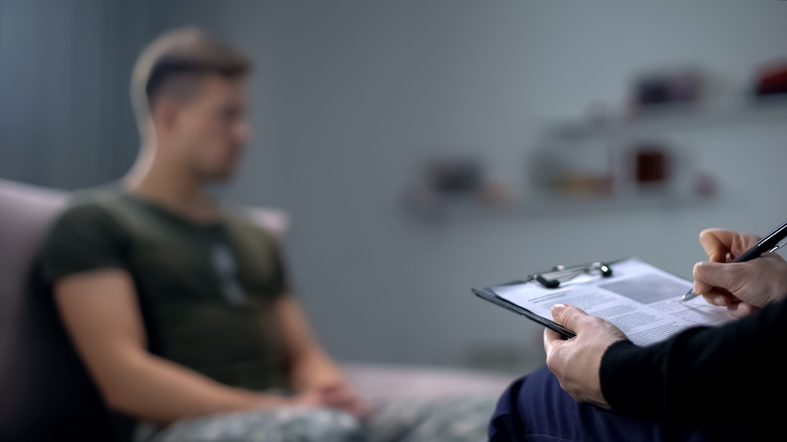 A person, seated and blurred in the background, appears to be having a discussion. In the foreground, another person holds a clipboard and pen, taking notes. The setting suggests a professional conversation likely taking place in a trauma clinic specializing in PTSD treatment.