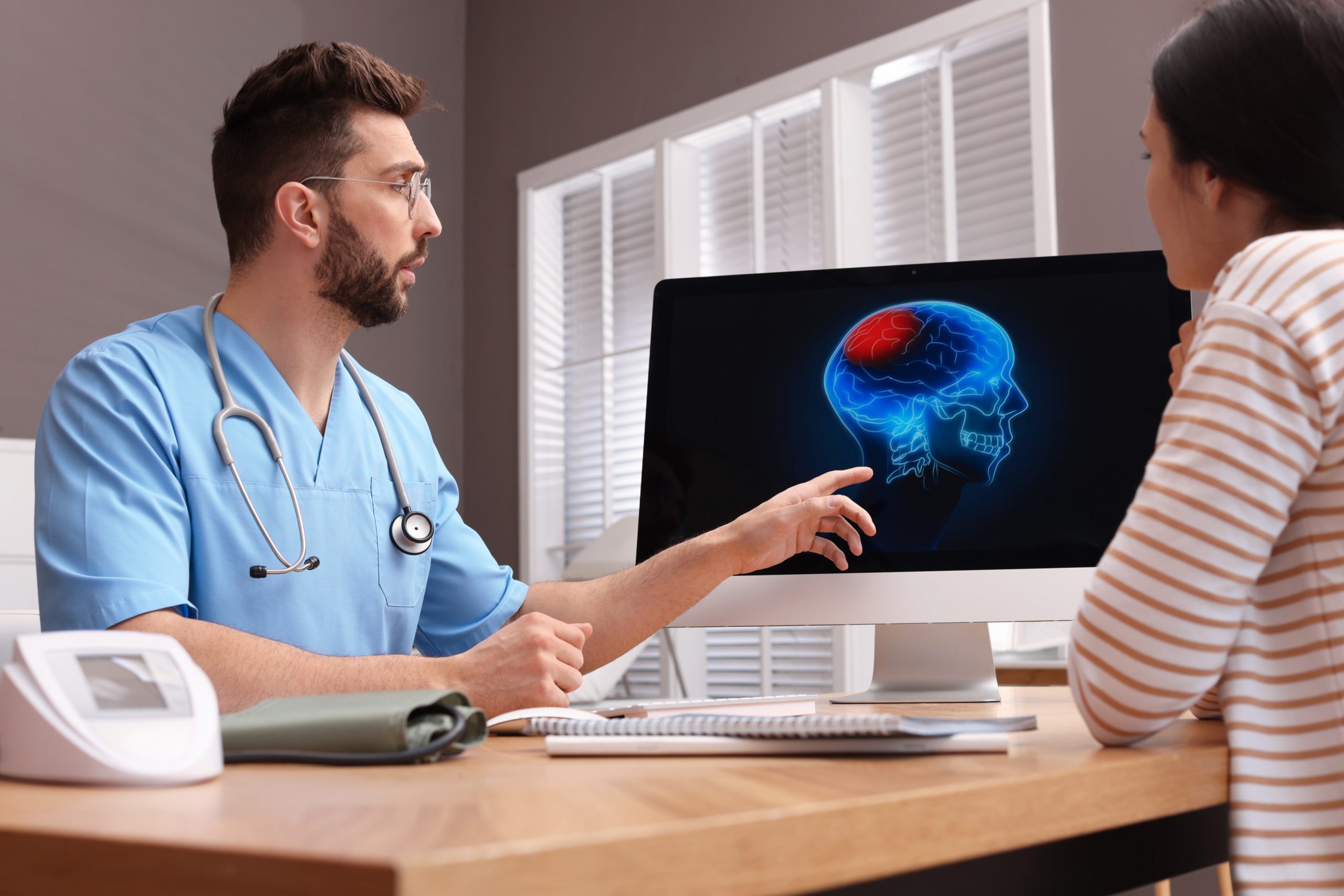 A male medical professional in blue scrubs, wearing a stethoscope, points at a computer screen displaying an image of a brain with a highlighted area in red. He is discussing PTSD treatment with a female patient seated across the desk in a striped shirt at Khiron Clinics.