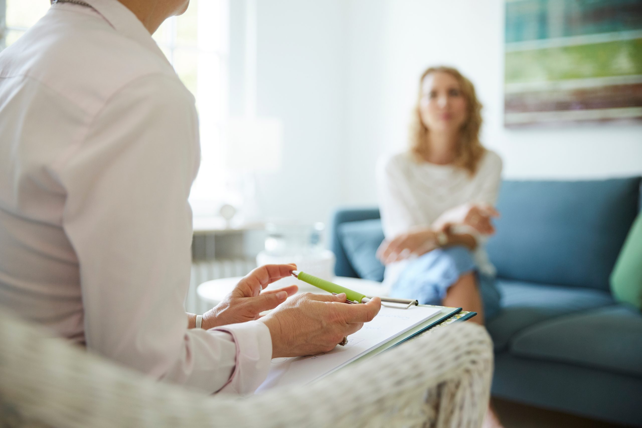 A therapist holding a clipboard and pen engages in discussion with a patient, who is sitting on a couch in a bright room. The patient appears relaxed and attentive, while the therapist is partially visible from the side, suggesting a professional mental health treatment setting.
