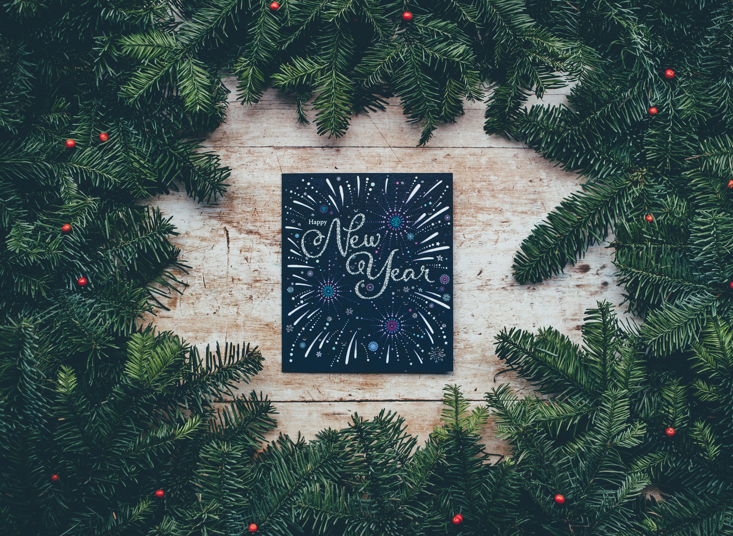 A black card with "Happy New Year" written in elegant cursive sits on a wooden surface, surrounded by a wreath of evergreen branches adorned with small red berries. The card features a firework design in the background, bringing warmth and hope akin to mental health treatment's uplifting spirit.