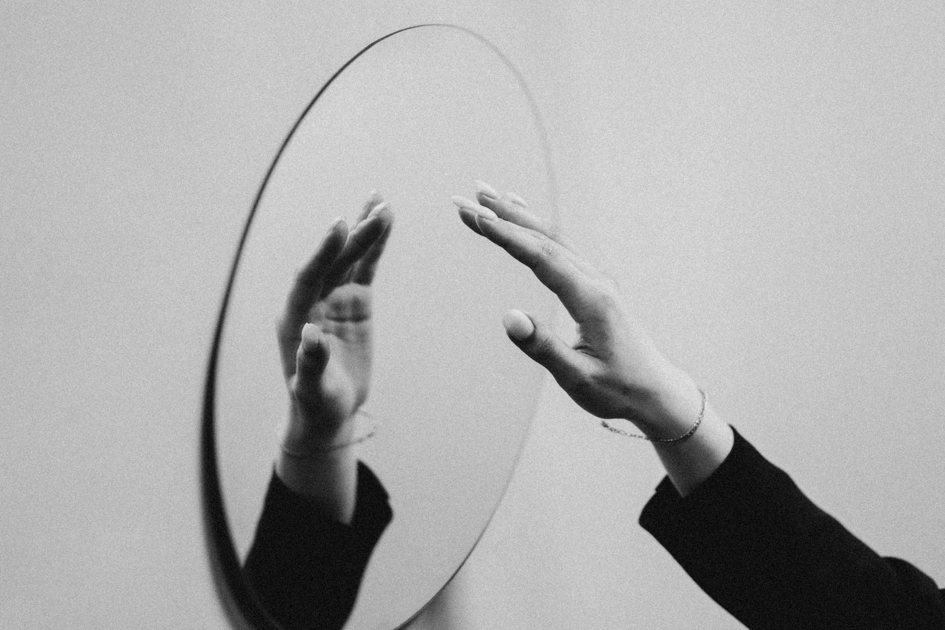 Grayscale image of a person's hand reaching towards a circular mirror, creating a reflection. The person appears to be wearing a dark long-sleeved garment and a thin bracelet on their wrist. The background is a plain, textured wall, evoking the introspection common in mental health treatment settings.