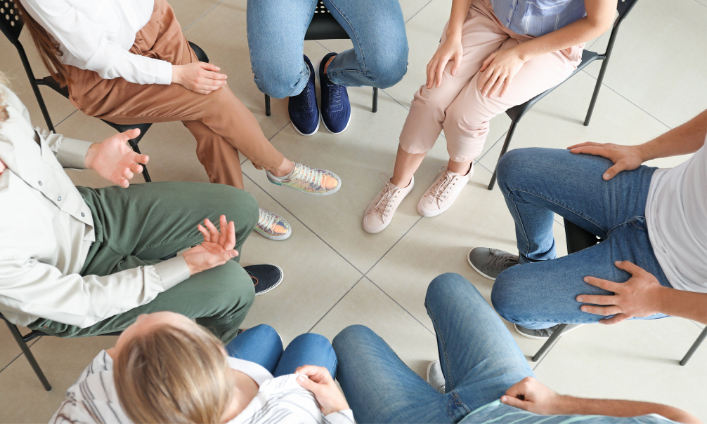 A group of people sitting in a circle on chairs. Their torsos and legs are visible, with various casual outfits including jeans, trousers, and sneakers. The setting appears to be an indoor space with a tiled floor at Khiron Clinics, suggesting a meeting or discussion focused on mental health.