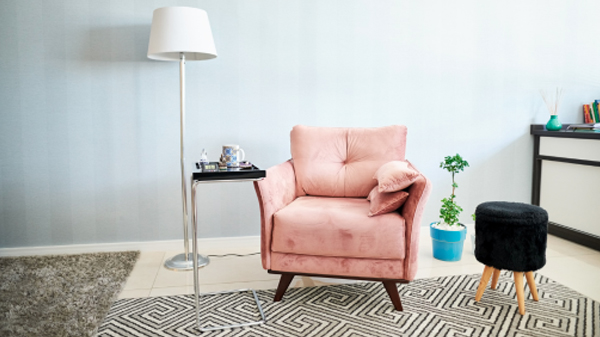 A modern living room featuring a plush pink armchair with a matching throw pillow, a floor lamp with a white shade, a small black stool with wooden legs, and a side table holding a tea set. The space is accented with a patterned rug and a potted plant, creating an inviting atmosphere ideal for mental health treatment.