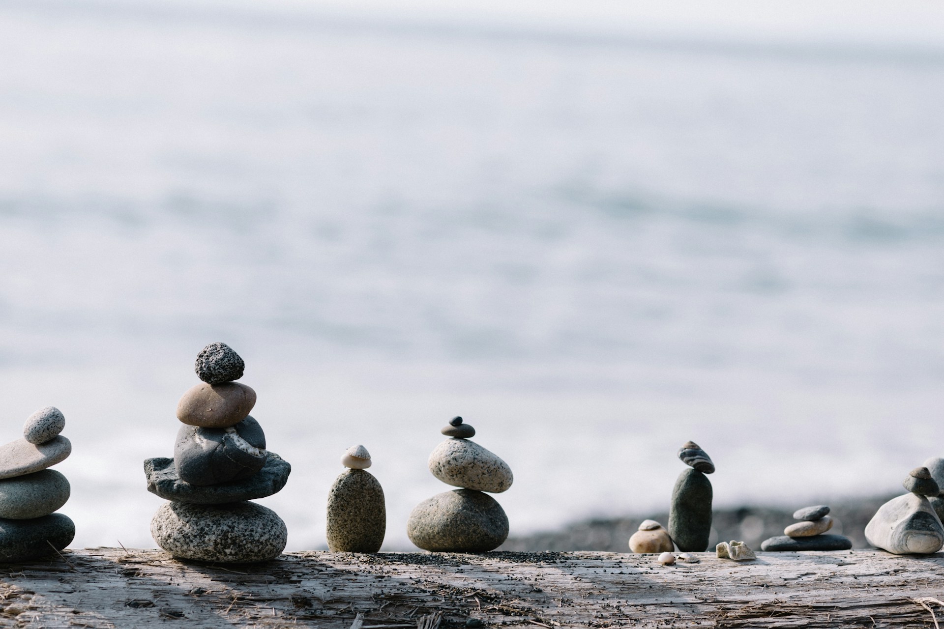 A collection of small stone stacks arranged on a driftwood log, with a blurred ocean in the background. The stacks vary in size and shape, reminiscent of the calming exercises used in trauma clinics to restore serenity and balance.