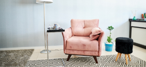 A cozy living room corner features a plush pink armchair with a small cushion, a modern floor lamp with a tray attached, a gray geometric rug, and a small black round stool. A potted plant sits in a blue pot near a black-and-white console table—an inviting space reminiscent of Khiron Clinics' soothing environments.