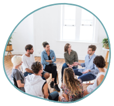A group of people sits in a circle in a well-lit room with wooden floors and potted plants. They appear to be engaged in a mental health treatment session or group therapy. Some individuals have their arms crossed, while others use hand gestures as they speak.