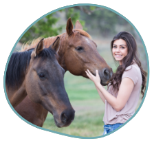 A woman with long dark hair is standing outdoors, smiling, and gently touching the face of a brown horse. Another brown horse is standing close by. The background, green with trees and grass, offers a serene setting that could aid in PTSD treatment - including equine therapy