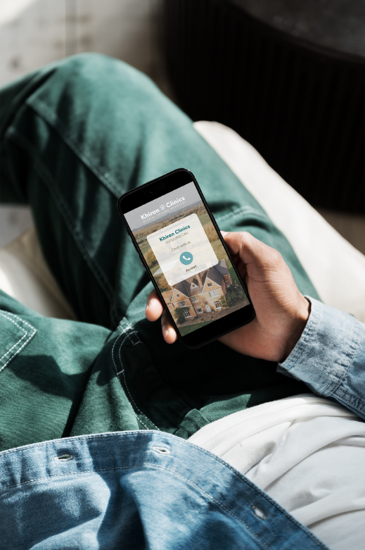 A person lounging on a couch uses a smartphone. The phone screen displays a vacation rental app showing details for a holiday cottage named “Russet Clines” with an image of the house and booking options. The person, wearing green pants and a denim shirt, appears to be unwinding after a session at Khiron Clinics.