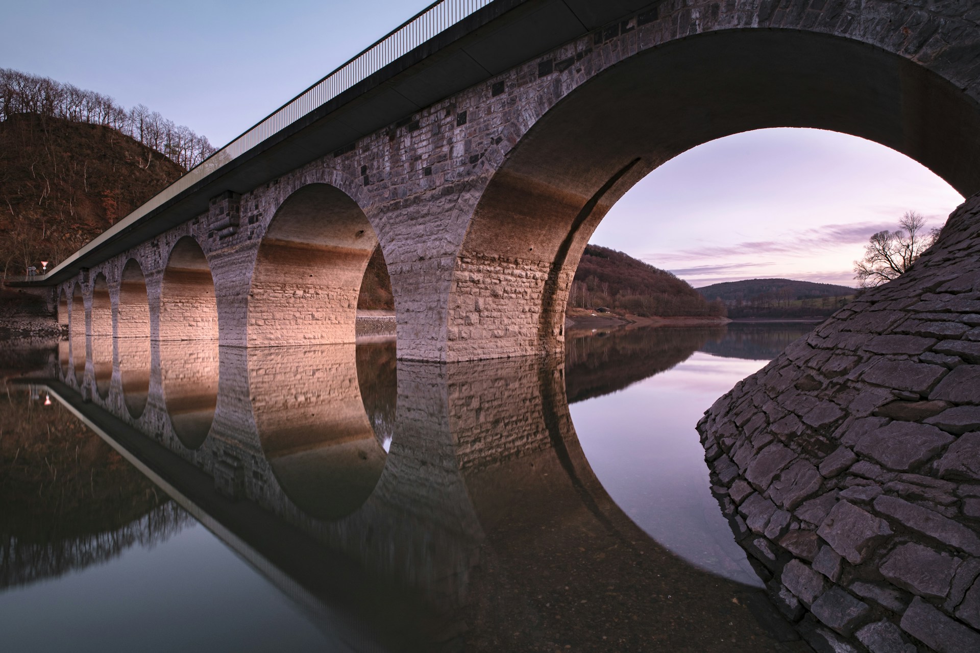 Stone bridge with multiple arches reflected in the calm water below. The landscape around the bridge is serene, with bare trees on the hills and a slightly pinkish sky at dusk. The overall atmosphere is tranquil and picturesque, offering a stark contrast to the origins of trauma hidden deep within nature’s quiet beauty.