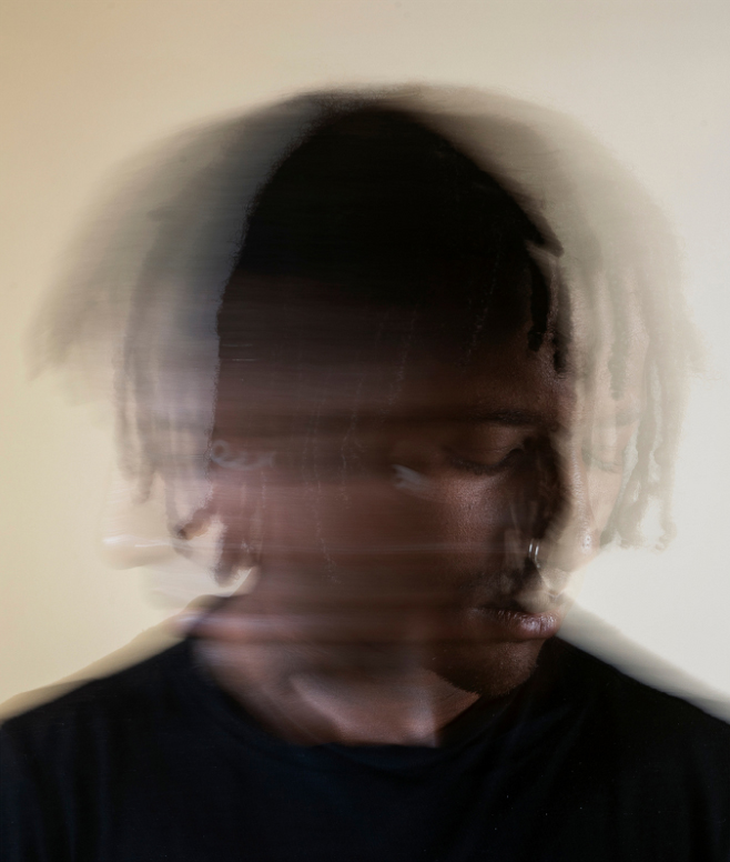 A blurred image of a person with medium to dark skin tone, wearing a black shirt. The motion blur effect creates an illusion of multiple faces overlaid, suggesting movement or rotation of the head. The background is neutral beige.