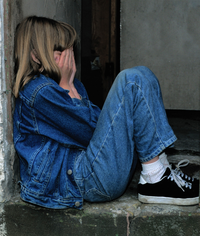 A young person, dressed in a denim jacket and jeans, sits with knees drawn up on a concrete step. They cover their face with both hands, appearing distressed, possibly contemplating mental health treatment. They wear black and white sneakers and white socks. The background includes a worn wall and a doorway.