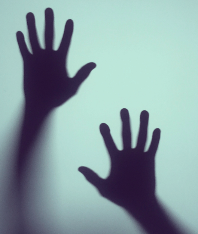 Two shadowy hand silhouettes, one larger and one smaller, are raised against a light background. The positioning suggests that the hands are pressing against or near a translucent surface, creating an eerie, almost ghostly effect—reminiscent of images often associated with trauma treatment settings like mental hospitals.