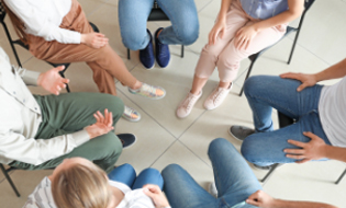 A group of people sit in a circle at Khiron Clinics, engaged in conversation or a meeting. They are seated on chairs with their legs and hands visible from an overhead perspective. The individuals are casually dressed in a variety of pants and shoes, participating in mental health treatment.