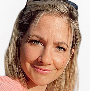 A woman with blonde hair smiles in a close-up portrait. She has bright eyes and wears a light pink top. Sunglasses rest on her head, and the plain white background highlights her features, similar to the welcoming atmosphere one can expect visiting About Khiron Clinics.