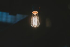 A single Edison-style light bulb is lit, emitting a warm glow in a dark and dimly lit environment. The filament inside the bulb is visible and slightly glowing, providing a soft, vintage ambiance that could soothe even the most frazzled nervous system.