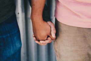 A close-up image of two people holding hands, with one person wearing a pink shirt and brown pants, and the other in a dark shirt and jeans. The background is blurred, revealing a textured, metallic surface. This tender moment captures the supportive bond often seen in trauma therapy sessions.