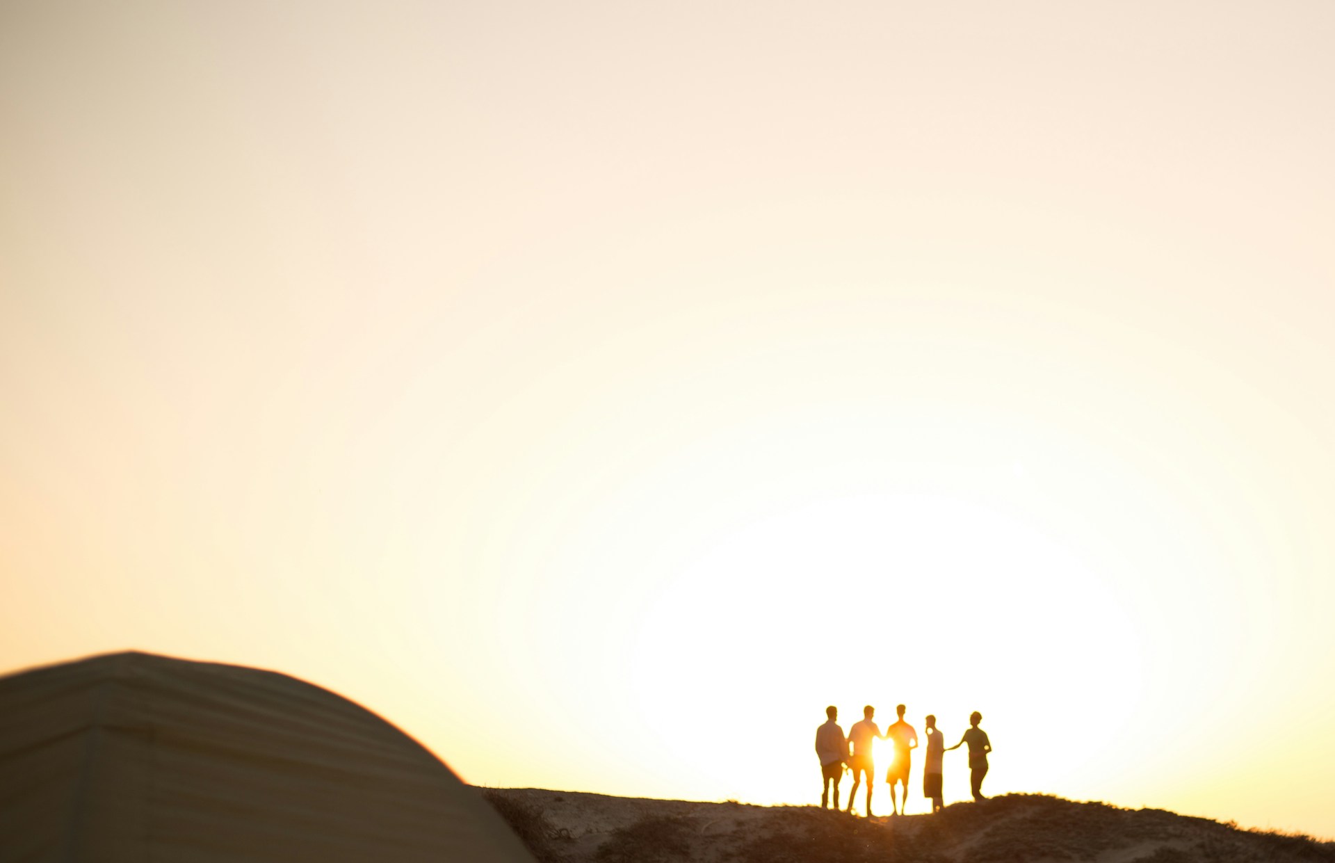 Silhouettes of five people stand on a hilltop against a bright, setting sun. The sky is a gradient of pale yellow to light orange. In the foreground, a structure with a domed roof is partially visible on the left side of the image, evoking thoughts about the risk of PTSD in such serene moments.