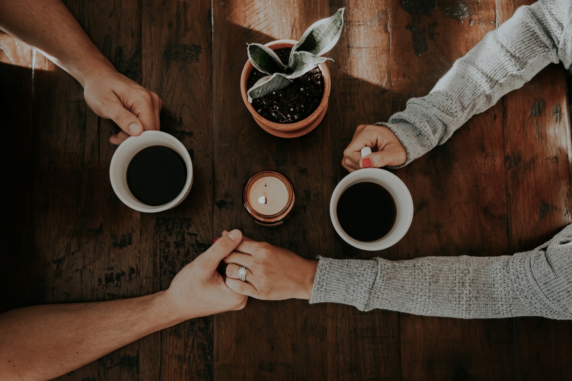 Two people are holding hands across a wooden table, each with a mug of coffee. A small plant and lit candle sit between them, creating a warm, cozy atmosphere as they support a loved one through conversation and connection.