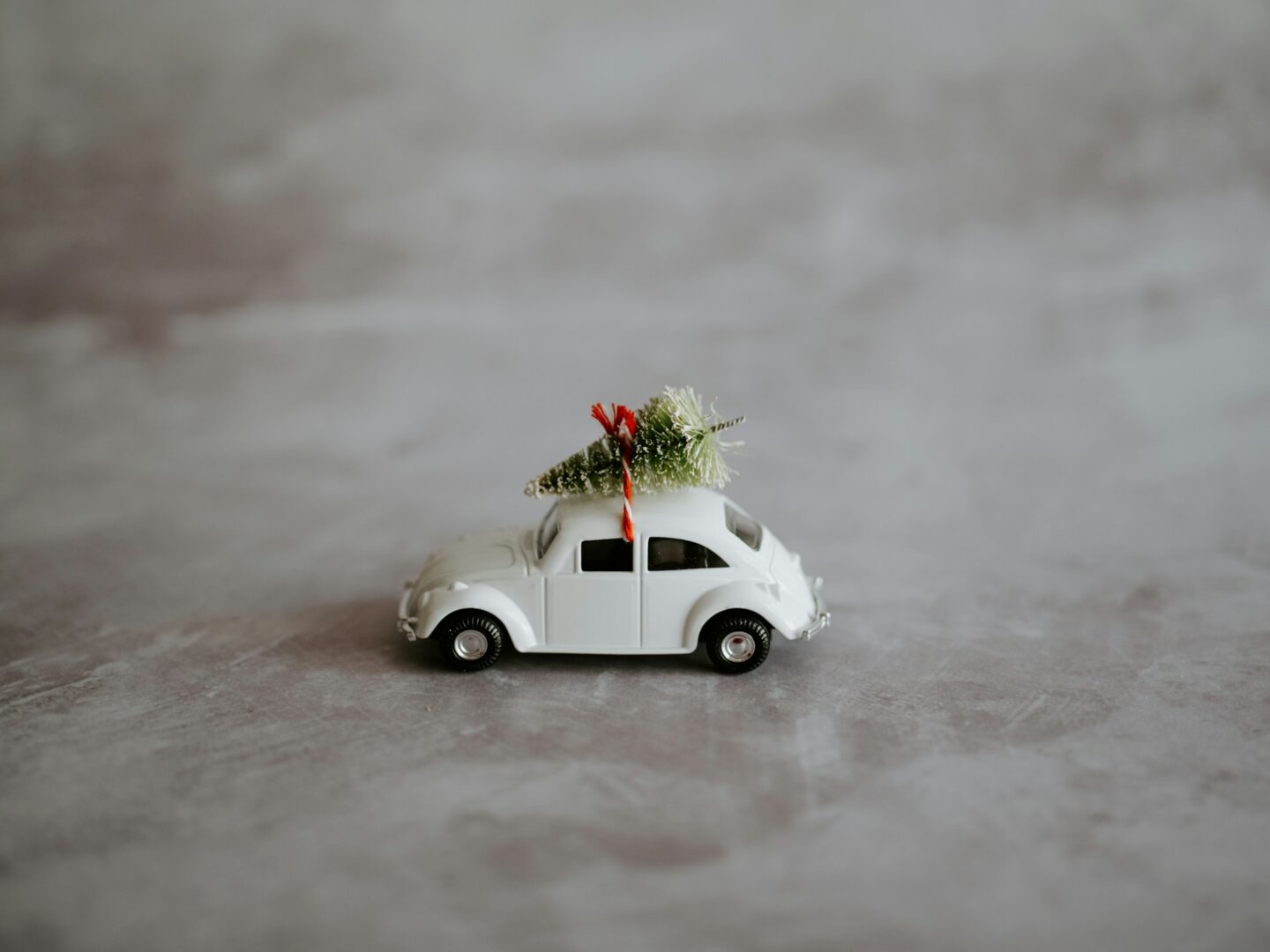 A vintage-style white toy car with a miniature Christmas tree tied on top with a red ribbon, placed on a textured gray surface, symbolizes resilience and peace during the holiday season.