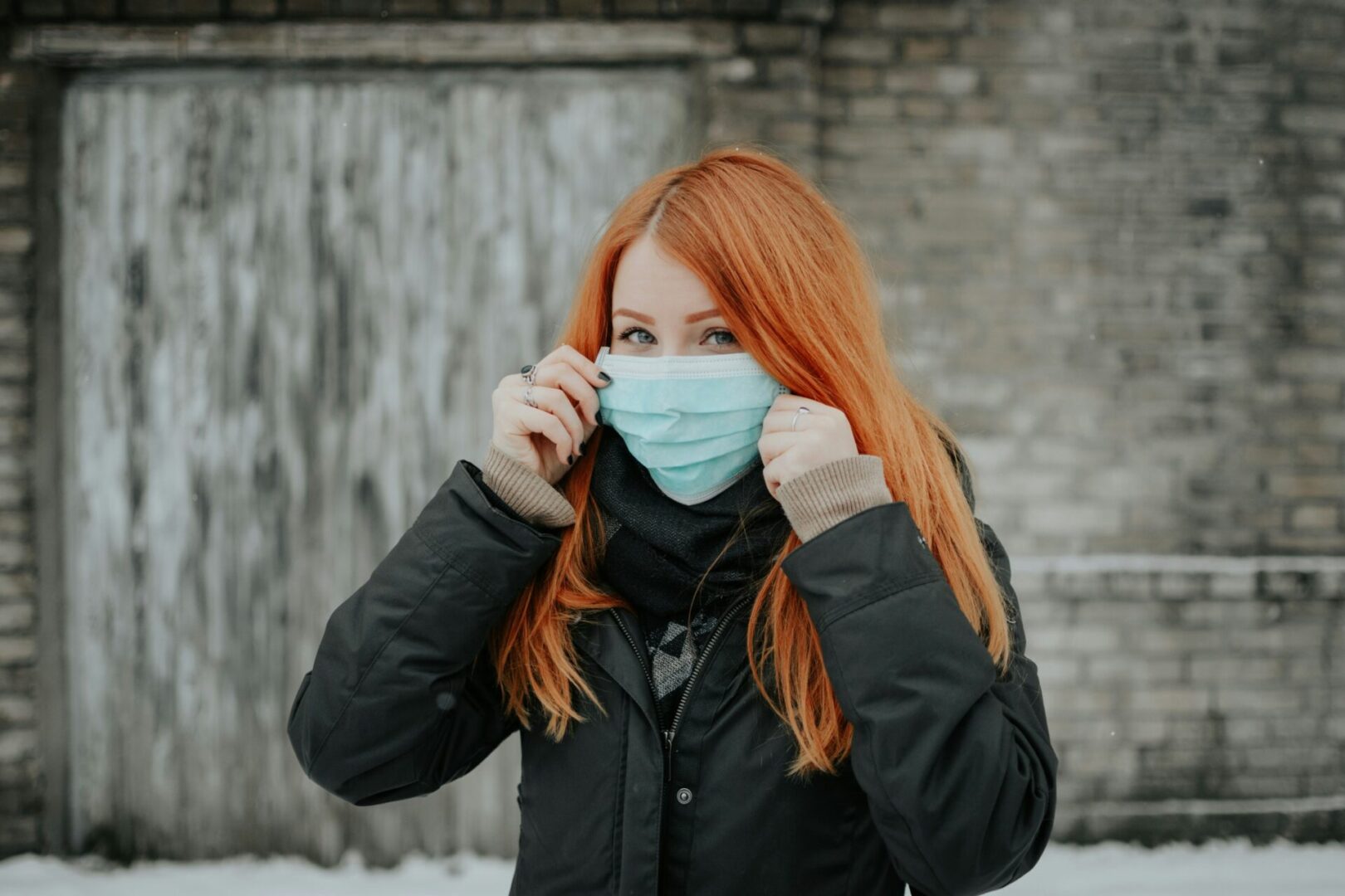 A person with long red hair adjusts a blue face mask while wearing a black coat and scarf. They stand outdoors in front of a weathered wooden door and a brick wall. The ground is covered with snow.