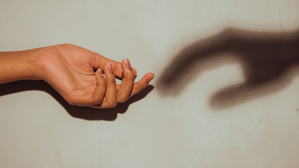 Close-up of a hand reaching out against a beige background, with a shadow of another hand mirroring its gesture on the wall. The image subtly hints at "What We Treat," emphasizing themes of connection and reaching out.