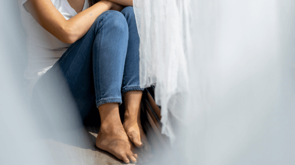 Person sitting by a window with sheer curtains, wearing a white shirt and blue jeans, barefoot. Wrapped in introspection and embodying the essence of "What We Treat," their arms encircle their knees, suggesting a contemplative mood.