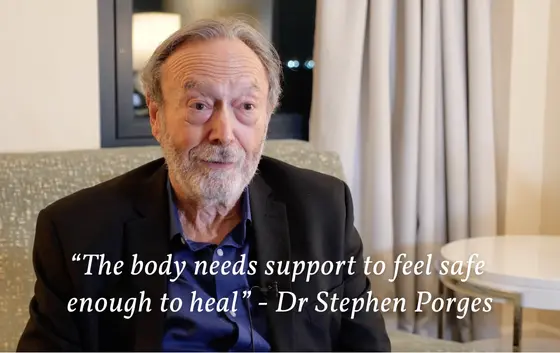 A man with a beard speaks indoors beside closed curtains, clad in a dark jacket over a blue shirt. Text on the image states, "The body needs support to feel safe enough to heal" — Dr. Stephen Porges. This insight resonates deeply in discussions on how we treat trauma today.