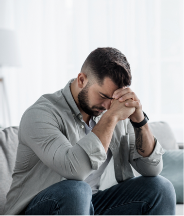 A man sits on a couch, leaning forward with his elbows on his knees and his hands clasped in front of his face.