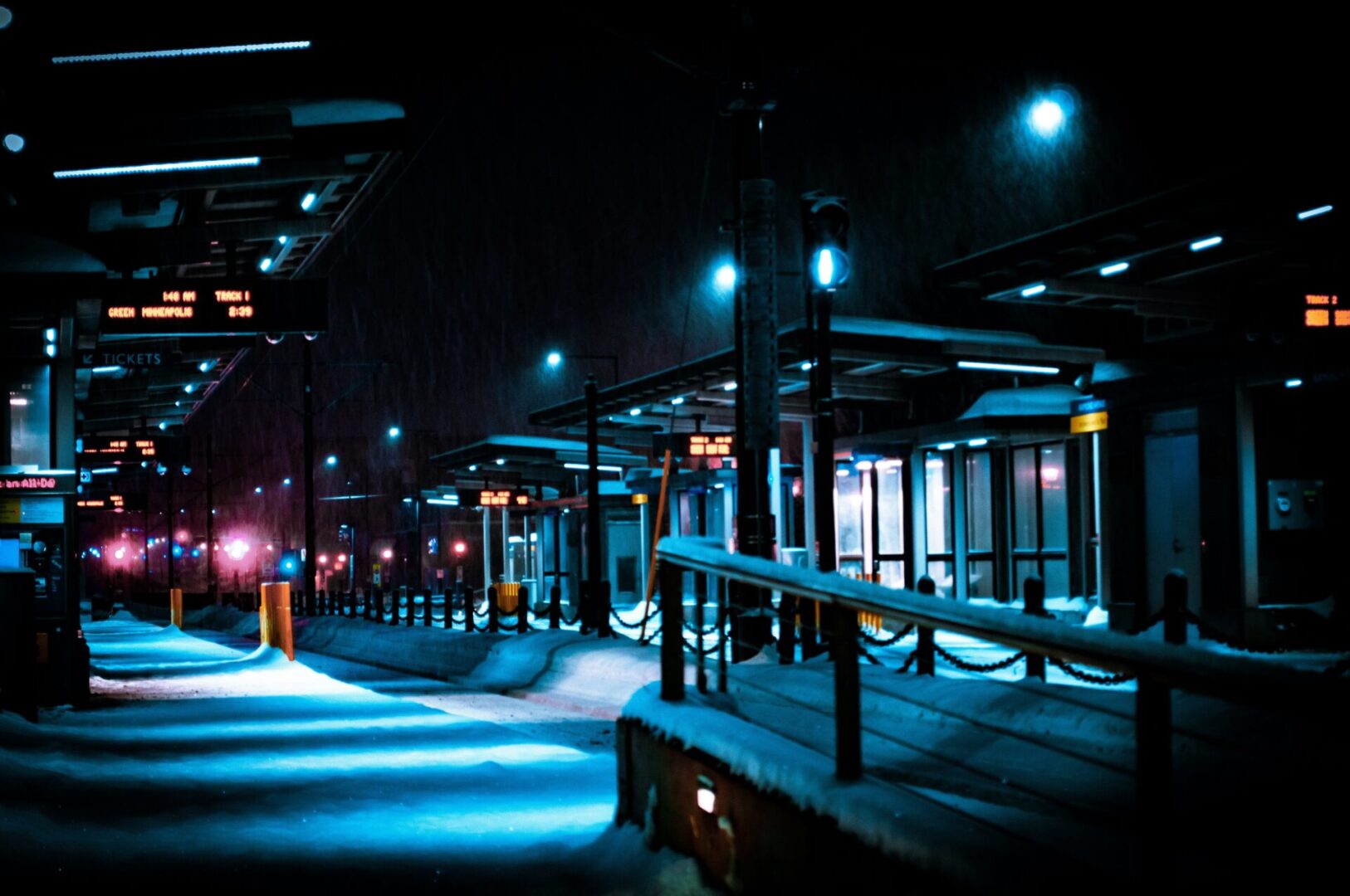 A snowy, deserted train station at night, illuminated by streetlights casting a bluish glow. The platform is layered in winter snow, and digital signs display information, creating a serene and quiet atmosphere.