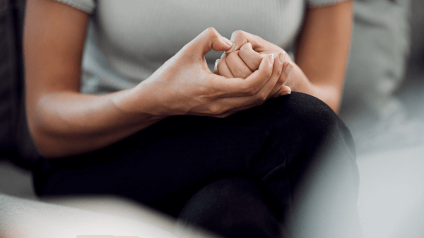 A person sits, hands resting in their lap, wearing a light gray shirt and black pants. Their intertwined fingers convey contemplation or anxiety, emblematic of the complex emotions addressed in What We Treat.