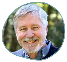 Smiling older man with gray hair and a beard, wearing a blue collared shirt and sweater, standing outdoors against a backdrop of greenery. His confident pose reflects his esteemed professional accreditations.