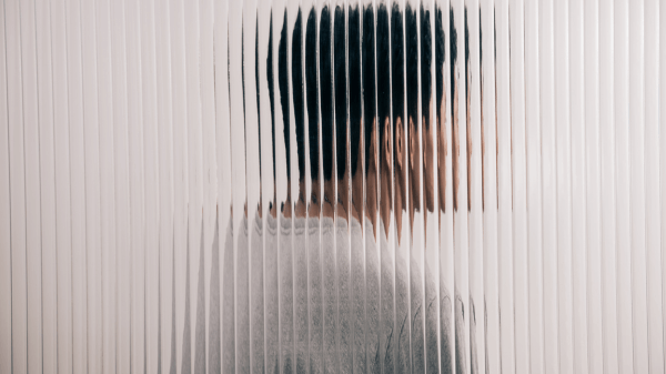 A person stands behind a textured glass panel, slightly blurred by the vertical lines. Facing to the right in a light-colored top, they have short, dark hair. This image subtly captures themes akin to the transformative clarity of "What We Treat," set against a neutral backdrop.