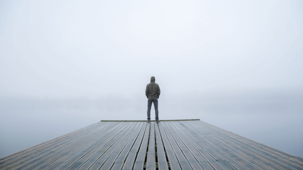 A person stands alone on a wooden platform, facing a foggy body of water. The overcast sky and mist create a serene and contemplative atmosphere, embodying the quiet introspection often found in what we treat. The person is wearing a hoodie and jeans.