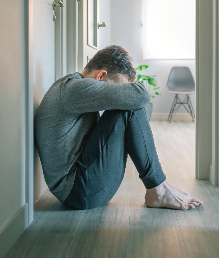 A person sits on the floor in a hallway, seeking stress relief as they lean against a wall with their head resting on crossed arms and knees drawn up. Wearing a gray long-sleeve shirt and dark pants, they relax as sunlight gently filters through a nearby window.