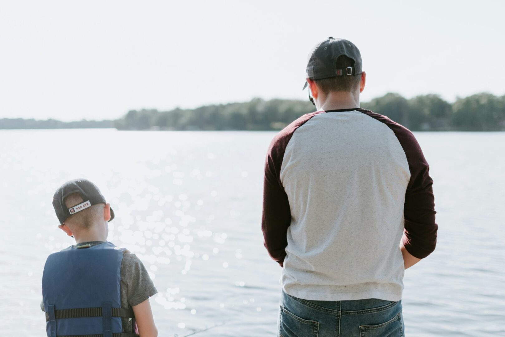 A man and a child stand by a calm lake, both wearing baseball caps. The child is in a blue life vest, as they engage in trauma-informed conversations while facing the water with trees in the distance. Sunlight sparkles on the serene surface, fostering an environment supporting children's mental health.