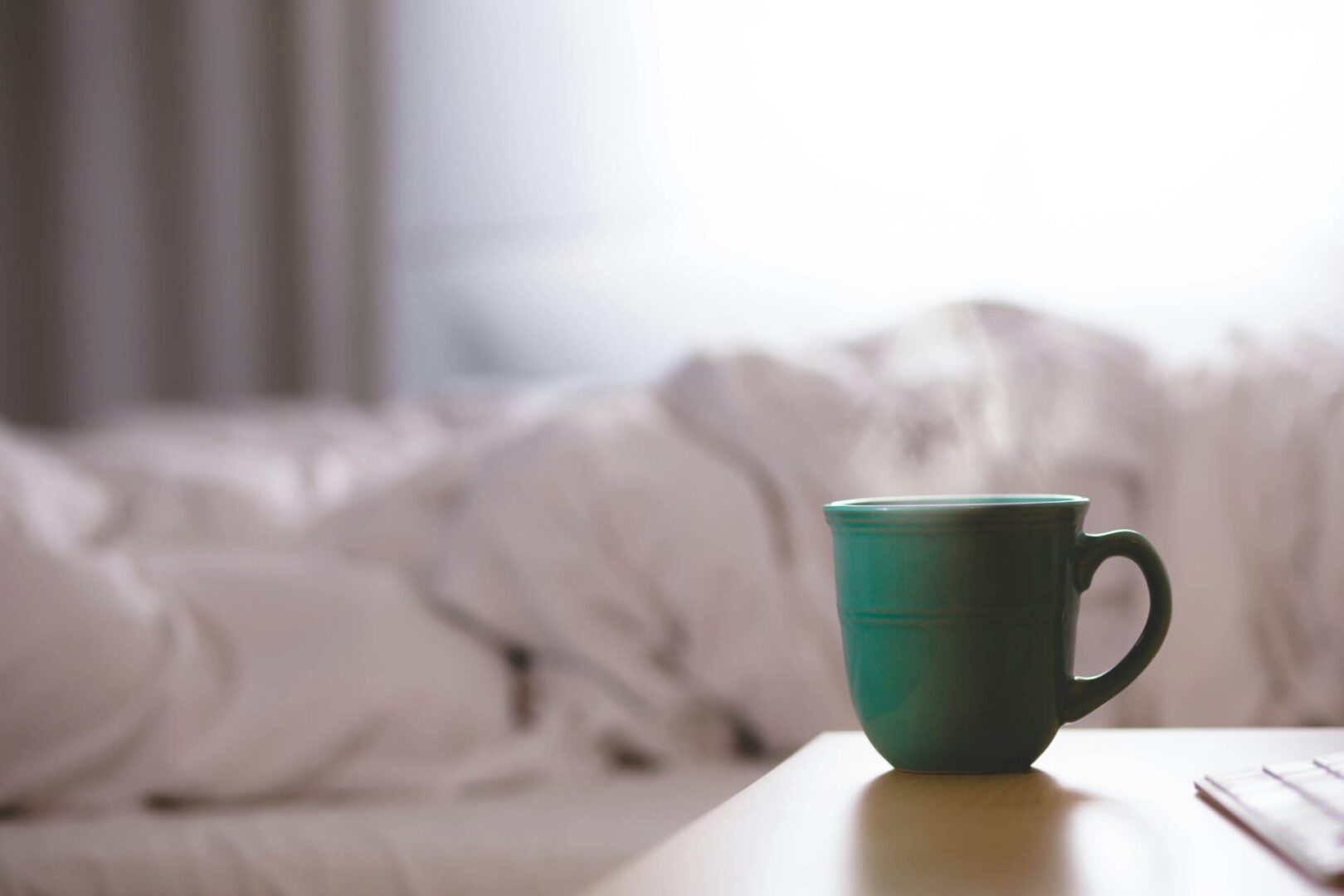 A green mug sits on a wooden table in the foreground, with a blurred, bright bedroom scene featuring unmade white bedding in the background. The cozy morning atmosphere contrasts with the remnants of nightmares lingering from sleep paralysis.