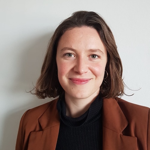A woman with short brown hair smiles at the camera. She is wearing a brown blazer over a black top and is standing in front of a plain white wall, reflecting the warm and professional approach found at About Khiron Clinics.