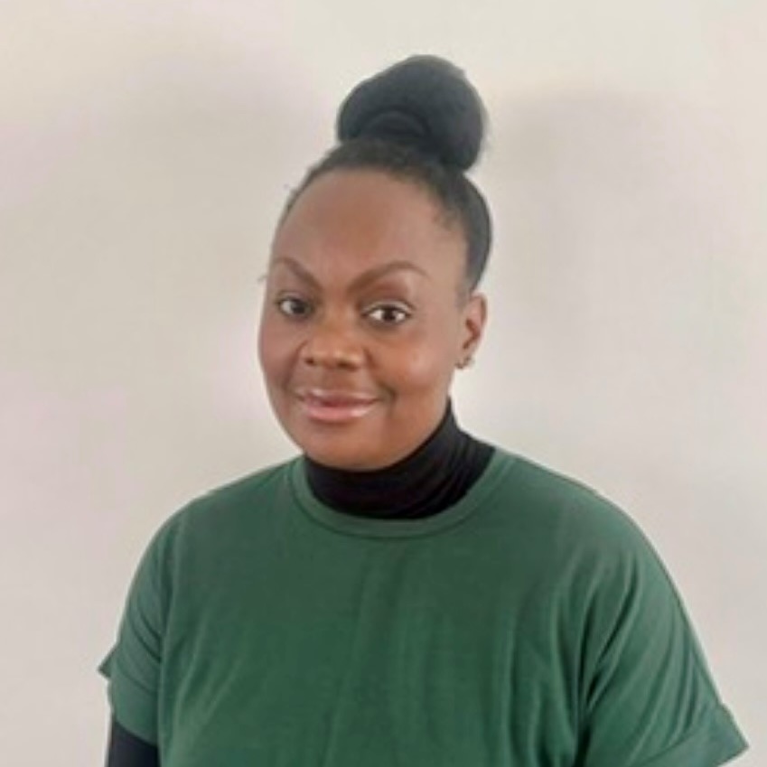 A woman with dark skin and hair in a high bun, wearing a green short-sleeve shirt over a black turtleneck, stands in front of a plain light-colored wall and smiles gently at the camera—reflecting the warmth found at Khiron Clinics.