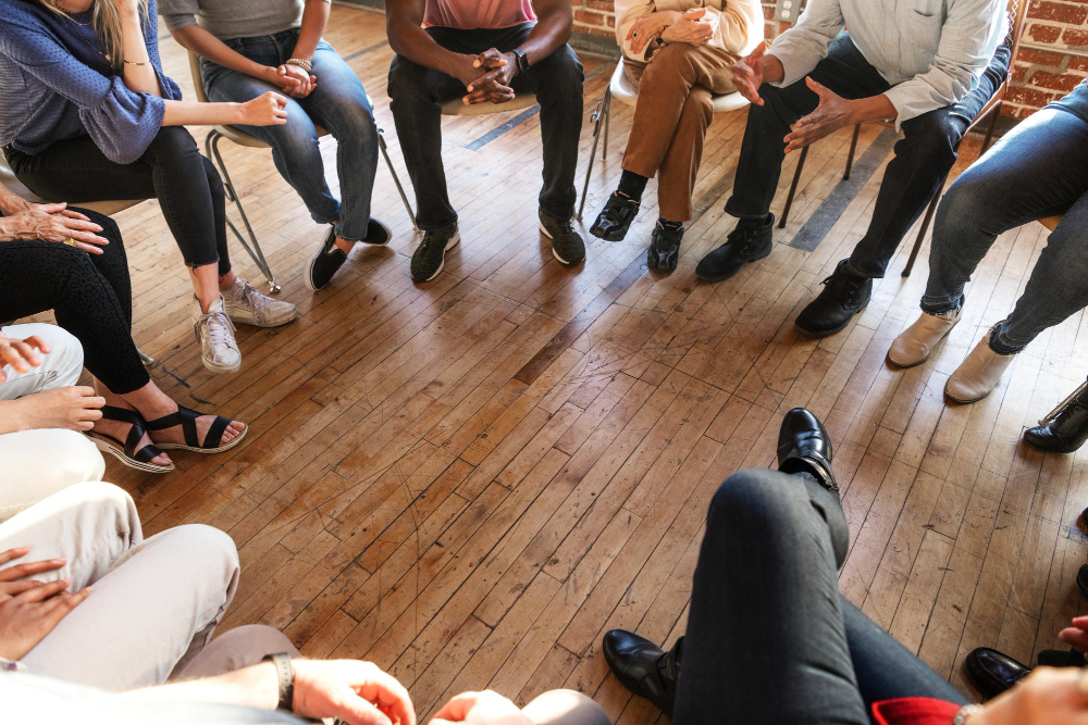A diverse group of people sits in a circle on chairs in a room with wooden floors, engaging in a group discussion focused on Trauma Recovery. Only their legs and hands are visible.