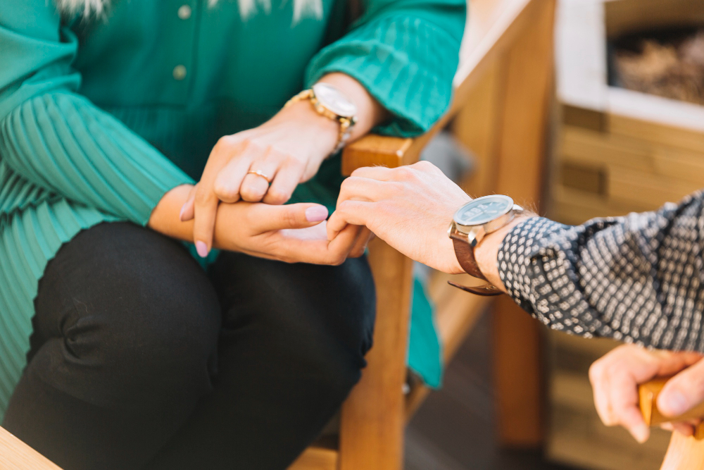 A close-up of two people holding hands. One, wearing a green top and watch, and the other in a checkered shirt, gently holds their fingers—capturing a moment of connection often seen in trauma recovery or nervous system-informed care in Oxfordshire.
