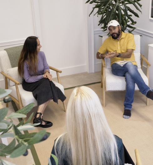Three people sit in armchairs having a conversation in a bright, modern room with plants. As one man gestures while discussing "What is Trauma," a woman listens intently and a third person with long blonde hair sits with their back to the camera.