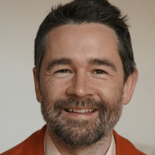 A man with short dark hair and a beard smiles at the camera. He is wearing a red sweater over a light-colored shirt, with a plain, light-toned background—reflecting the warm, welcoming atmosphere found at About Khiron Clinics.