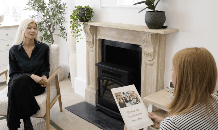 Two women sit in a bright, cozy room at the Trauma Clinic. One holds a booklet titled "Guide to Treatment" while the other listens attentively, suggesting a thoughtful discussion during a therapy or counseling session.