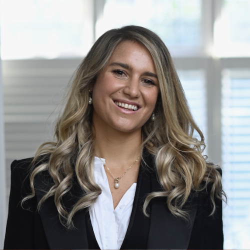 A woman with long, wavy blonde hair wearing a black blazer and white blouse smiles at the camera. She stands indoors with sunlight coming through white blinds, reflecting the warm, welcoming atmosphere often found at Khiron Clinics.