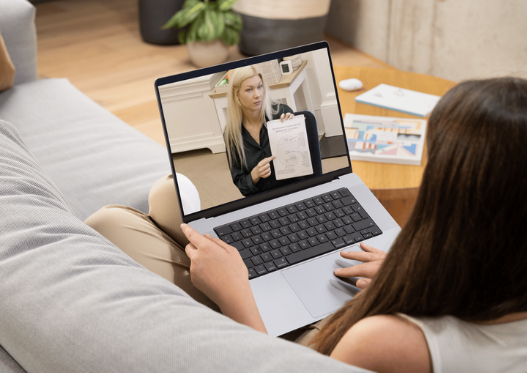 A person sitting on a couch uses a laptop for a video call about What is Trauma, as another woman on screen shows paperwork and explains something. A notebook and pen are on the coffee table nearby.