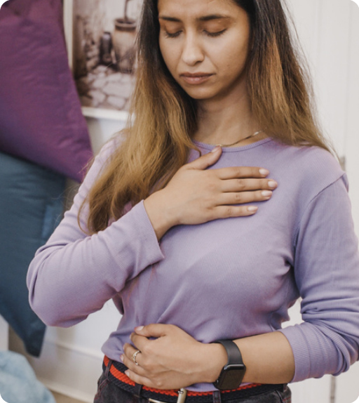A woman wearing a purple top holds one hand on her chest and the other on her stomach, eyes closed, as if focusing on her breathing—capturing a moment that may prompt the question: What is trauma?.