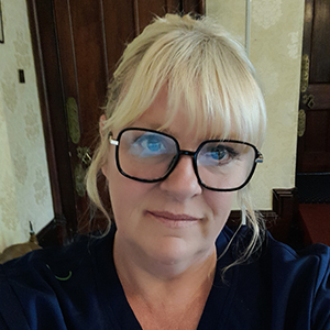 A woman with blonde hair and bangs, wearing large black glasses and a dark top, stands indoors in front of wooden doors and patterned wallpaper—an atmosphere reminiscent of the welcoming environment at Khiron Clinics.